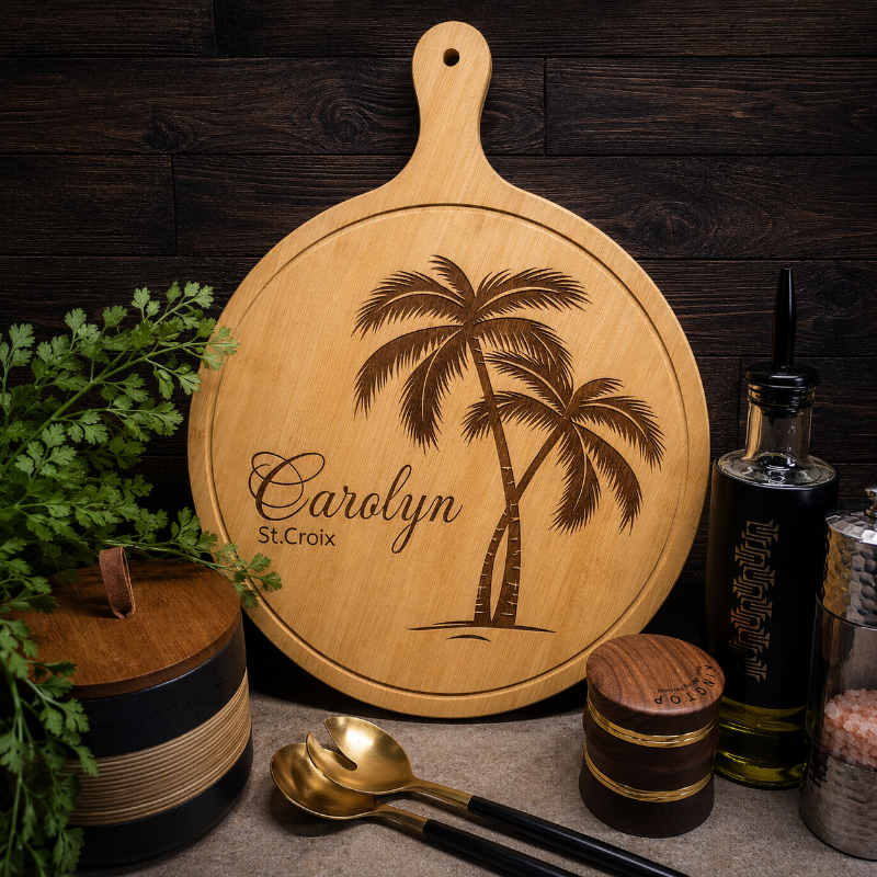 Wooden cutting board with palm tree design and engraved name, surrounded by kitchen items on a wooden surface.