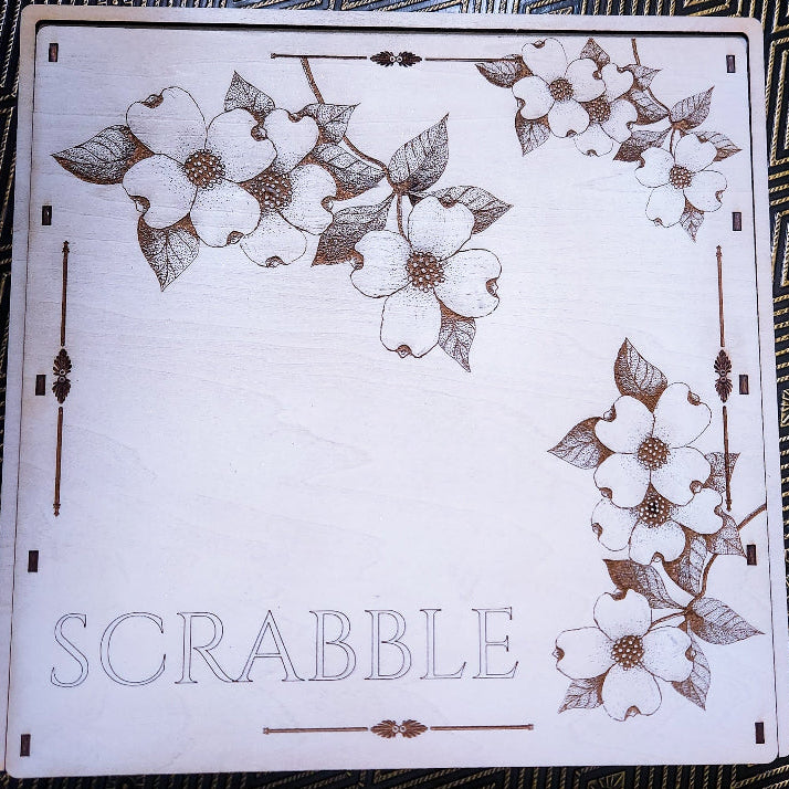 A square white wooden Scrabble board with engraved brown floral designs and the word 'SCRABBLE' at the bottom, photographed from above on a dark patterned fabric background.