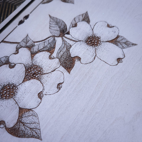 A close-up of a white wood surface featuring a detailed brown etched or burned floral design with five-petal blossoms and leaves.