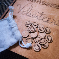 A close-up of engraved wooden football-shaped tokens or coins arranged on a tan leather-like surface next to a small white drawstring pouch. The surface shows partial engraved text including 'TENNESSE' and 'Volunteers'.
