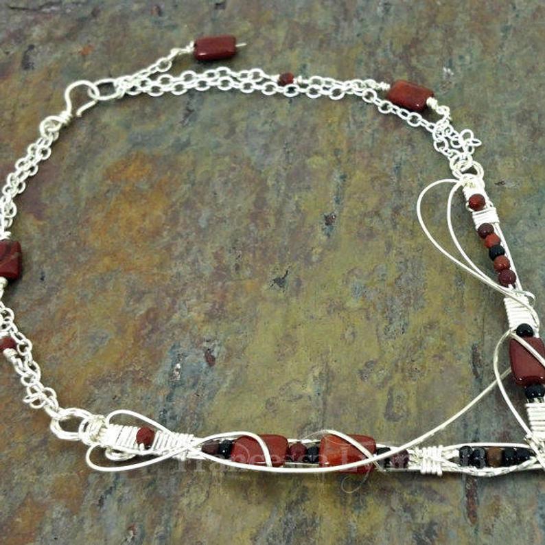 This collar is hand crafted and wrapped with strands of luscious sterling silver wire and embellished with Red Jasper and Black Onyx. Gemstones and accent beads are incorporated securely into the wire work. Flowing curves soften the hard lines of the metals and an asymmetrical flair makes this an unusual and dramatic addition to your private collection.